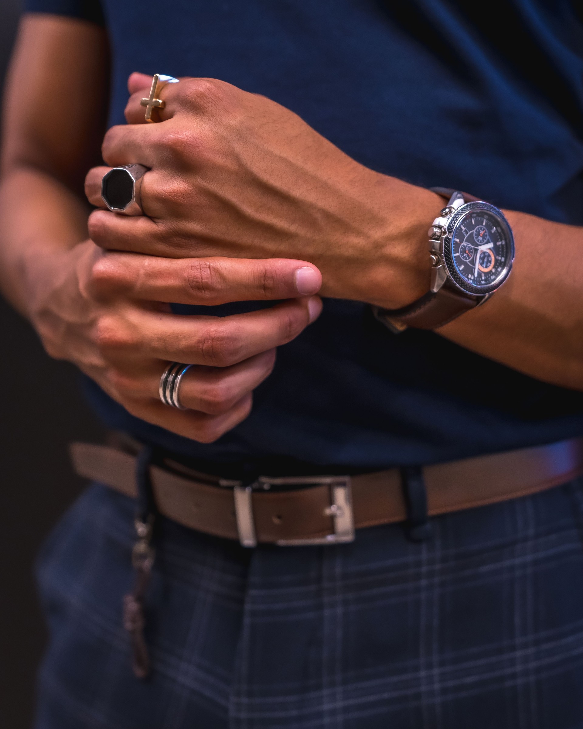 Fashion lifestyle, portrait of a young Latino man on a black wall. Blue polo shirt and plaid pants. Detail of hands with rings and watch